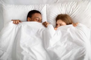 A man and woman peeking at each other under the covers of a bed.
