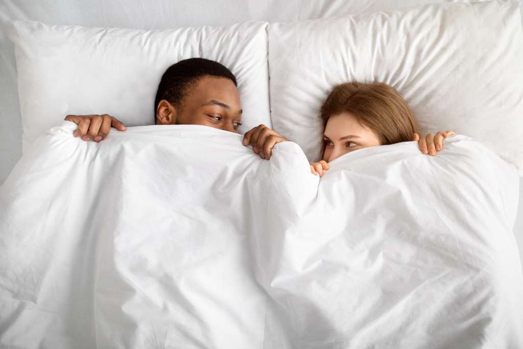 A man and woman peeking at each other under the covers of a bed.